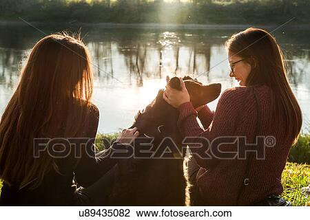 Two young women petting dog on river bank, Calolziocorte, Lombardy, Italy View Large Photo Image Stock Image - Two young women petting dog on river bank, Calolziocorte, Lombardy, Italy. Fotosearch