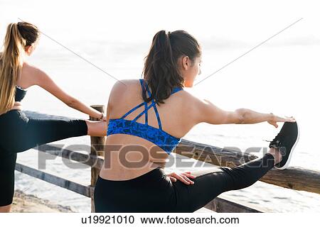 Stock Image - Two young women training on sea waterfront, touching toes. Fotosearch