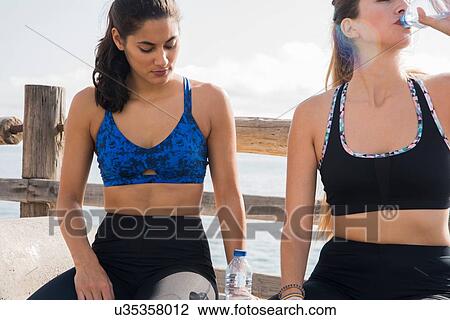 Stock Image - Two young women training together on sea waterfront, drinking bottled water. Fotosearch