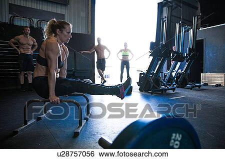 Stock Photograph - Woman exercising in gymnasium, using parallel bars, in L-sit position, friends in background, watching. Fotosearch