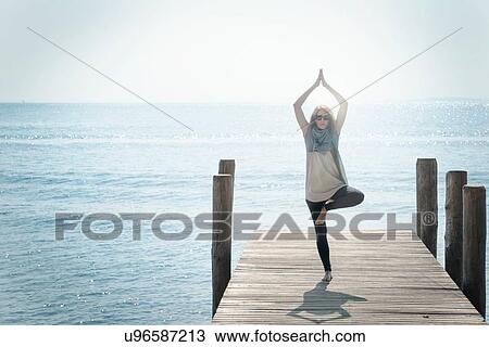 Stock Image - Woman on pier balancing on one leg in yoga pose. Fotosearch