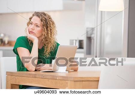 Stock Image - Woman sitting at kitchen table, laptop in front of her, thoughtful expression. Fotosearch