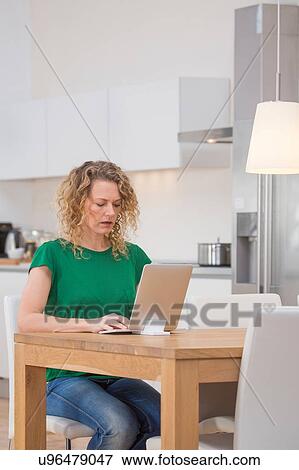 Stock Photo - Woman sitting at kitchen table, using laptop. Fotosearch