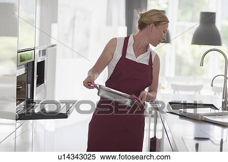 Stock Photography - Woman standing with hot baking pan and checking recipe. Fotosearch