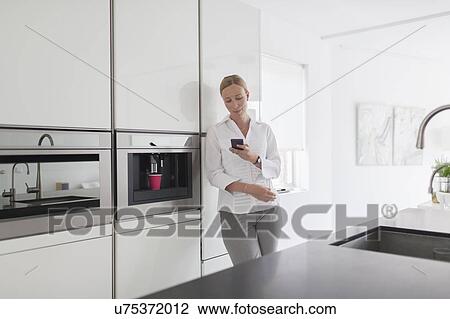 Stock Image - Woman using phone in kitchen. Fotosearch
