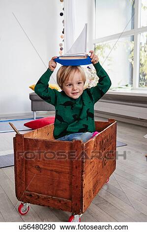 Stock Image - Young boy sitting in toy box, holding toy boat on head. Fotosearch