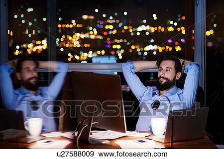 Stock Photo - Young businessman at office desk looking at computer at night, mirror image. Fotosearch