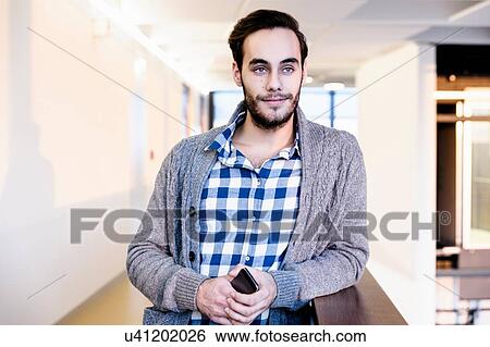 Stock Photograph - Young businessman holding smartphone in office corridor. Fotosearch
