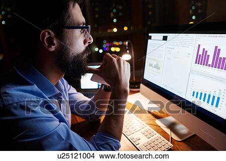 Picture - Young businessman looking at computer on office desk at night. Fotosearch