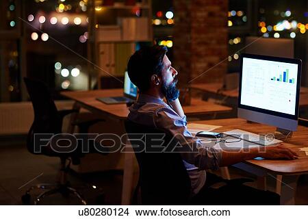 Picture - Young businessman looking at computer on office desk at night. Fotosearch
