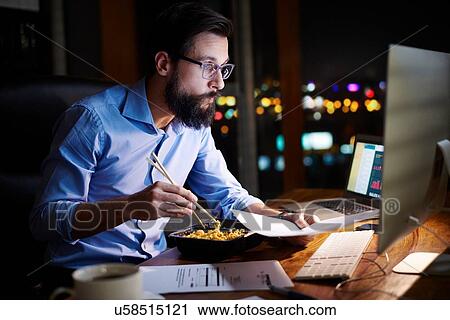 Stock Image - Young businessman looking at computer and eating takeaway at office desk at night. Fotosearch