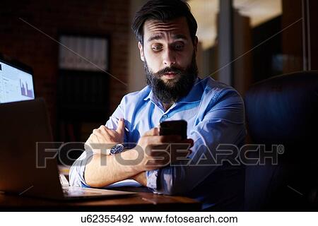 Young businessman looking at smartphone at office desk at night View Large Photo Image Stock Image - Young businessman looking at smartphone at office desk at night. Fotosearch
