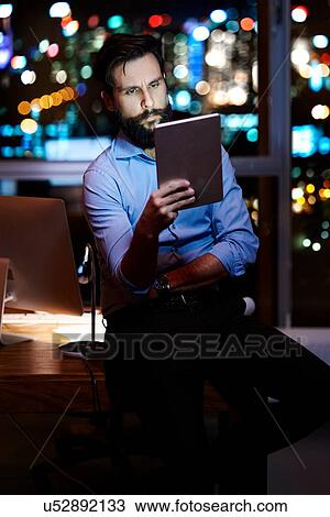 Stock Image - Young businessman on office desk looking at digital tablet at night. Fotosearch