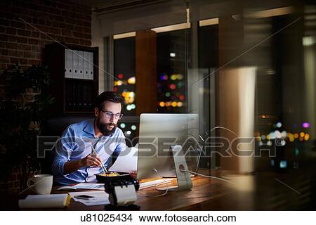 Young businessman reading paperwork and eating takeaway at office desk at night View Large Photo Image Picture - Young businessman reading paperwork and eating takeaway at office desk at night. Fotosearch