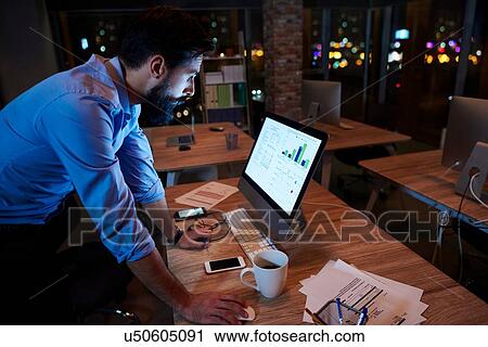 Stock Image - Young businessman working at computer on office desk at night. Fotosearch