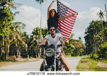 Stock Photo - Young couple holding up American flag while riding motorcycle on rural road, Krabi, Thailand. Fotosearch