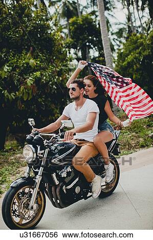Stock Photograph - Young couple holding up American flag while riding motorcycle on rural road, Krabi, Thailand. Fotosearch