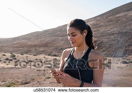Picture - Young female runner looking at smartphone in arid landscape, Las Palmas, Canary Islands, Spain. Fotosearch
