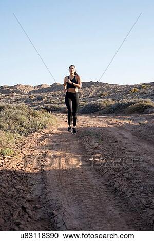 Stock Image - Young female runner running along dirt track in arid landscape, Las Palmas, Canary Islands, Spain. Fotosearch