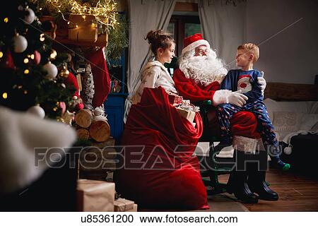 Stock Image - Young girl and boy sitting with Santa, sack full of presents beside him. Fotosearch