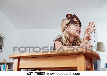 Stock Image - Young girl sitting at table, wearing bear mask, low angle view. Fotosearch