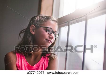 Stock Image - Young girl, wearing glasses, looking out of window. Fotosearch