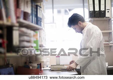 Stock Image - Young male pharmacist weighing pill bottle on pharmacy scales. Fotosearch