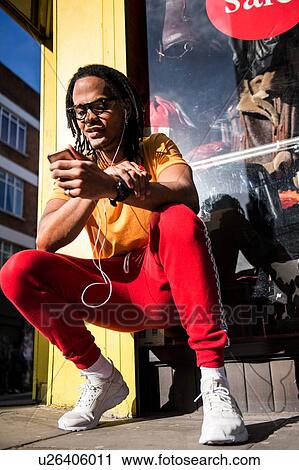 Young man crouching outdoors, wearing earphones, using smartphone View Large Photo Image Stock Image - Young man crouching outdoors, wearing earphones, using smartphone. Fotosearch