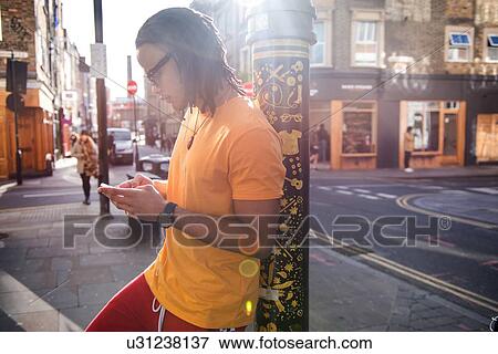 Stock Photo - Young man in street, leaning against lamppost, using smartphone. Fotosearch