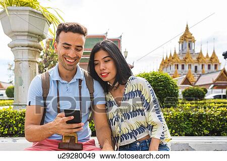 Stock Photo - Young tourist couple looking at smartphone outside temple, Bangkok, Thailand. Fotosearch