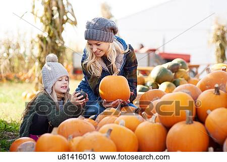 Stock Image - Young woman and daughter selecting pumpkin from stack at pumpkin patch. Fotosearch