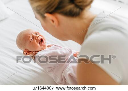 Young woman cradling crying baby daughter on bed, over shoulder view View Large Photo Image Stock Photo - Young woman cradling crying baby daughter on bed, over shoulder view. Fotosearch