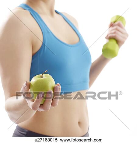 Stock Photography - Young woman holding apple and hand weight, studio shot.. Fotosearch