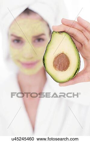 Stock Image - Young woman holding half an avocado wearing face mask, studio shot.. Fotosearch