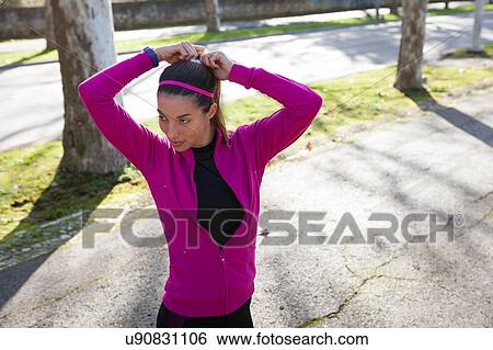 Stock Photograph - Young woman in street tying hair in ponytail. Fotosearch