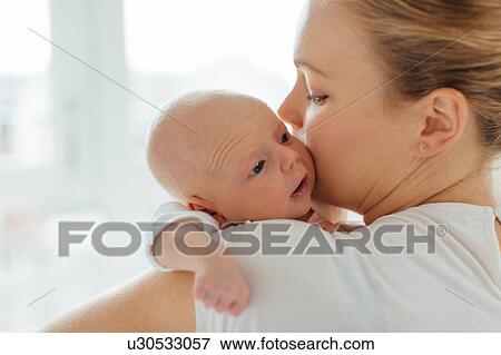 Stock Photo - Young woman kissing baby daughter on cheek, over shoulder view. Fotosearch