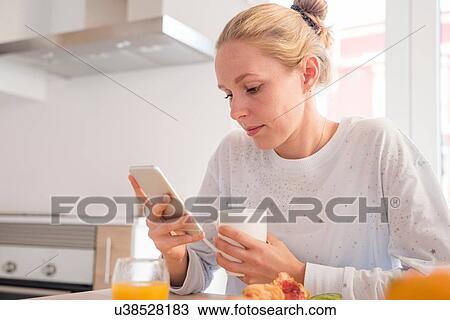 Stock Image - Young woman looking at smartphone at breakfast table. Fotosearch