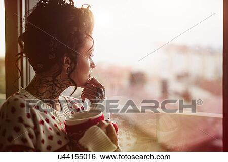 Stock Photograph - Young woman looking out of window, holding hot drink, thoughtful expression. Fotosearch