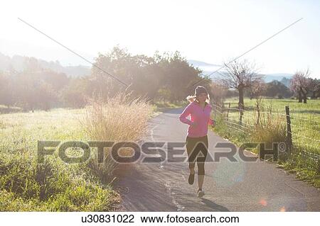Stock Image - Young woman running along rural road. Fotosearch