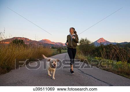 Stock Photo - Young woman running along rural road with pet dog. Fotosearch
