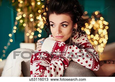 Stock Image - Young woman sitting at home, wearing christmas jumper, thoughtful expression. Fotosearch