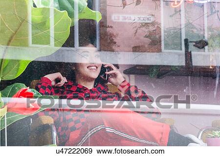 Stock Photo - Young woman sitting indoors, using smartphone, viewed through window. Fotosearch