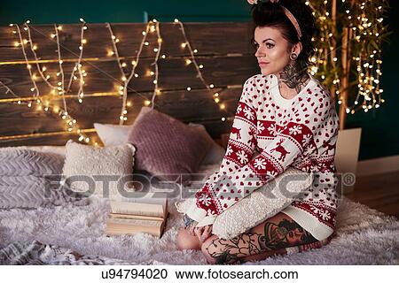 Stock Image - Young woman sitting on bed, wearing christmas jumper, thoughtful expression. Fotosearch