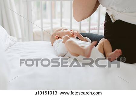 Stock Image - Young woman sitting on bed with baby daughter, cropped. Fotosearch