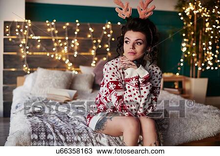 Young woman sitting on edge of bed, wearing christmas jumper and antlers, thoughtful expression View Large Photo Image Stock Image - Young woman sitting on edge of bed, wearing christmas jumper and antlers, thoughtful expression. Fotosearch