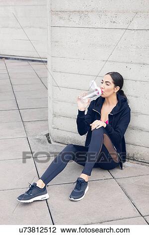 Stock Image - Young woman sitting on floor drinking water.. Fotosearch