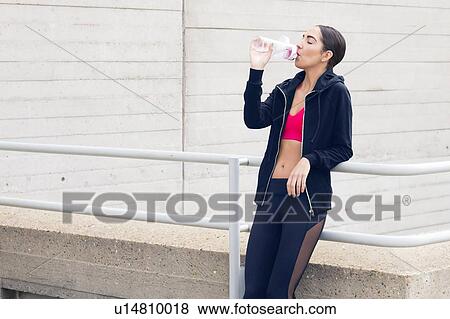 Stock Photo - Young woman standing by building drinking water.. Fotosearch