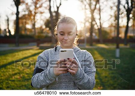 Stock Image - Young woman training in park looking at smartphone. Fotosearch