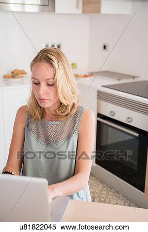 Stock Photography - Young woman typing on laptop at kitchen table. Fotosearch