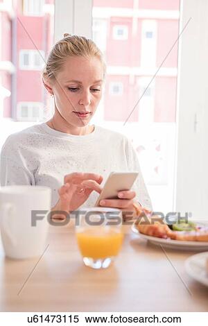 Stock Photography - Young woman using smartphone touchscreen at breakfast table. Fotosearch